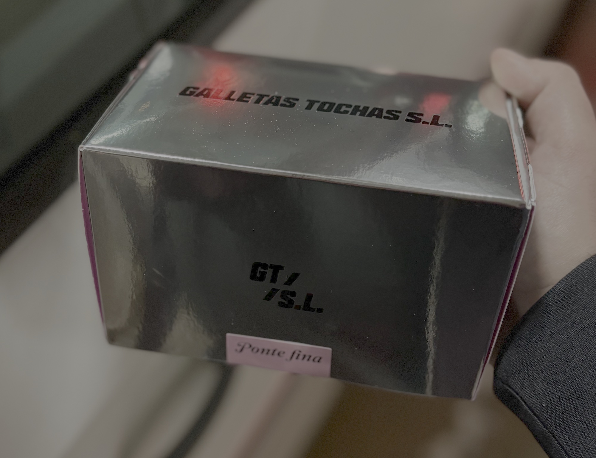 Caja metalizada de Galletas Tochas SL con una etiqueta rosa que reza "Ponte fina". El ambiente es oscuro y con luces reflejándose en al caja. Podría ser nu parking.