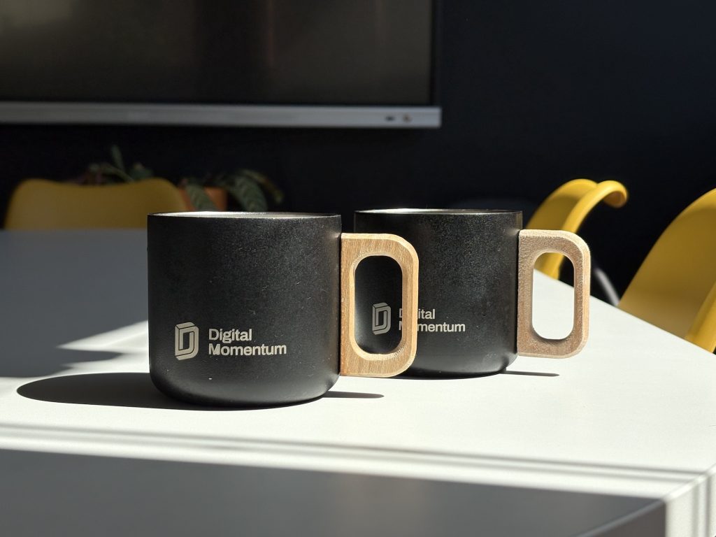Dos tazas de café metálicas con asa de madera y la marca "Digital Momentum" en una sala de reuniones soleada