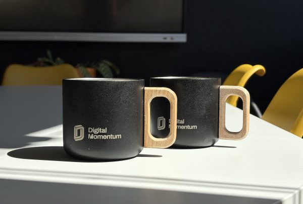 Dos tazas de café metálicas con asa de madera y la marca "Digital Momentum" en una sala de reuniones soleada